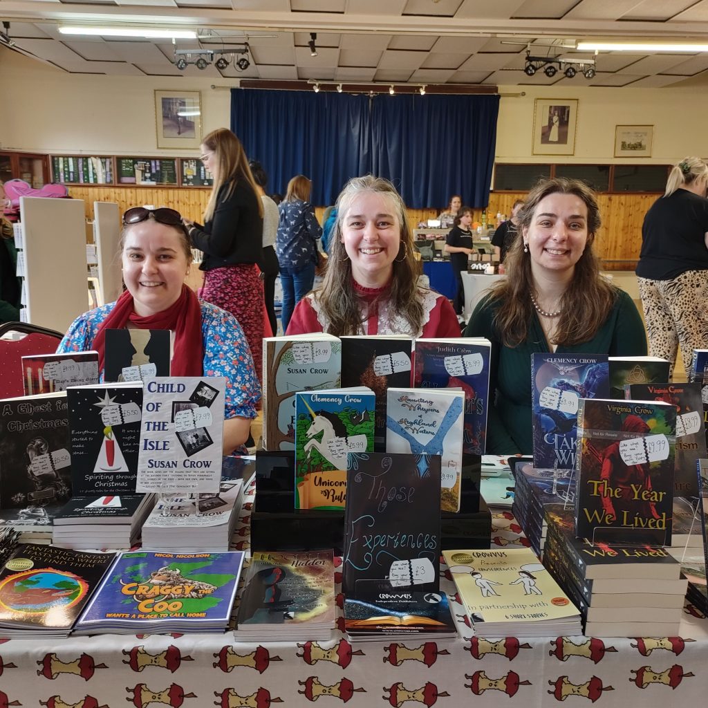 A book stall table at a local craft fayre. Three women sit cheerfully behind the table, smiling for the camera.
