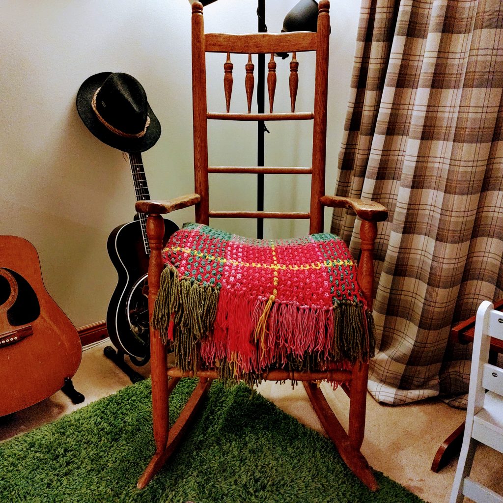 A colourful knitted blanket is folded up on an old-fashioned rocking chair. In the background are two guitars.