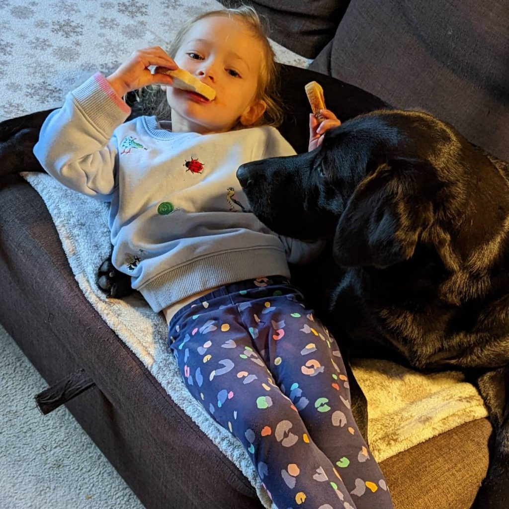 A pre-school girl leans back against a black dog, eating some toast and looking directly at the camera. The dog's head is curved round to watch the toast carefully.