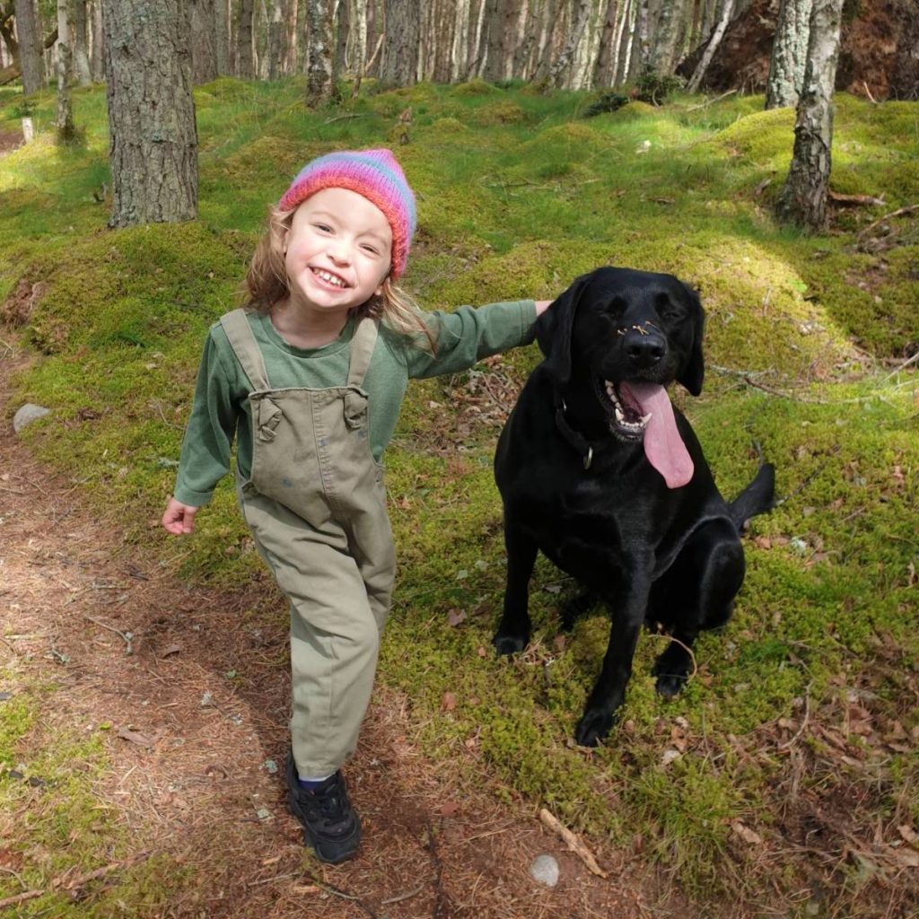 A young pre-school girl stands grinning next to her black dog in a wood, her hand on the dog's head. She is wearing green dungarees and a striped woolly hat. The dog has needles from the trees on her nose, and her ridiculously long tongue is hanging out of her mouth.