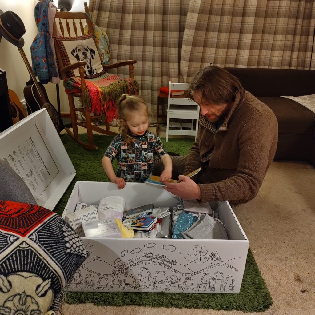 A Baby Box, full of clothes and other things needed for a brand new baby, sits in the middle of a room. A preschooler and her dad are looking through the contents and reading the books together.