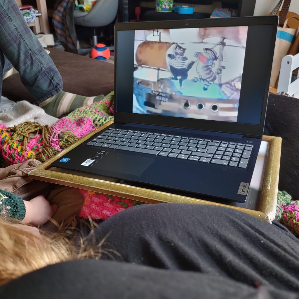 The programme Bagpuss is showing on a laptop screen. At the corners of the image can be seen the back of a preschooler's head as she watches the programme, her parents, and a black dog sprawled asleep on the settee. 