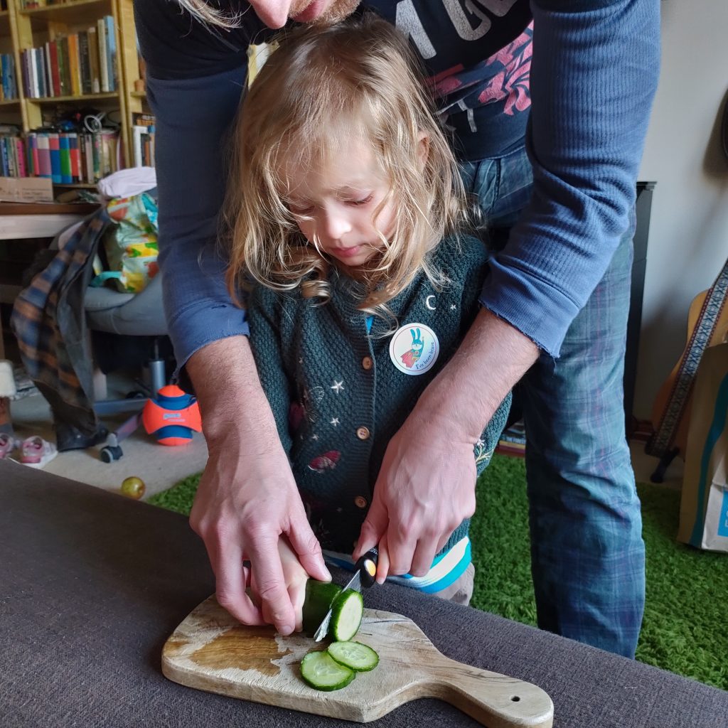 A preschooler chops a cucumber using a safety knife, with her father's help.
