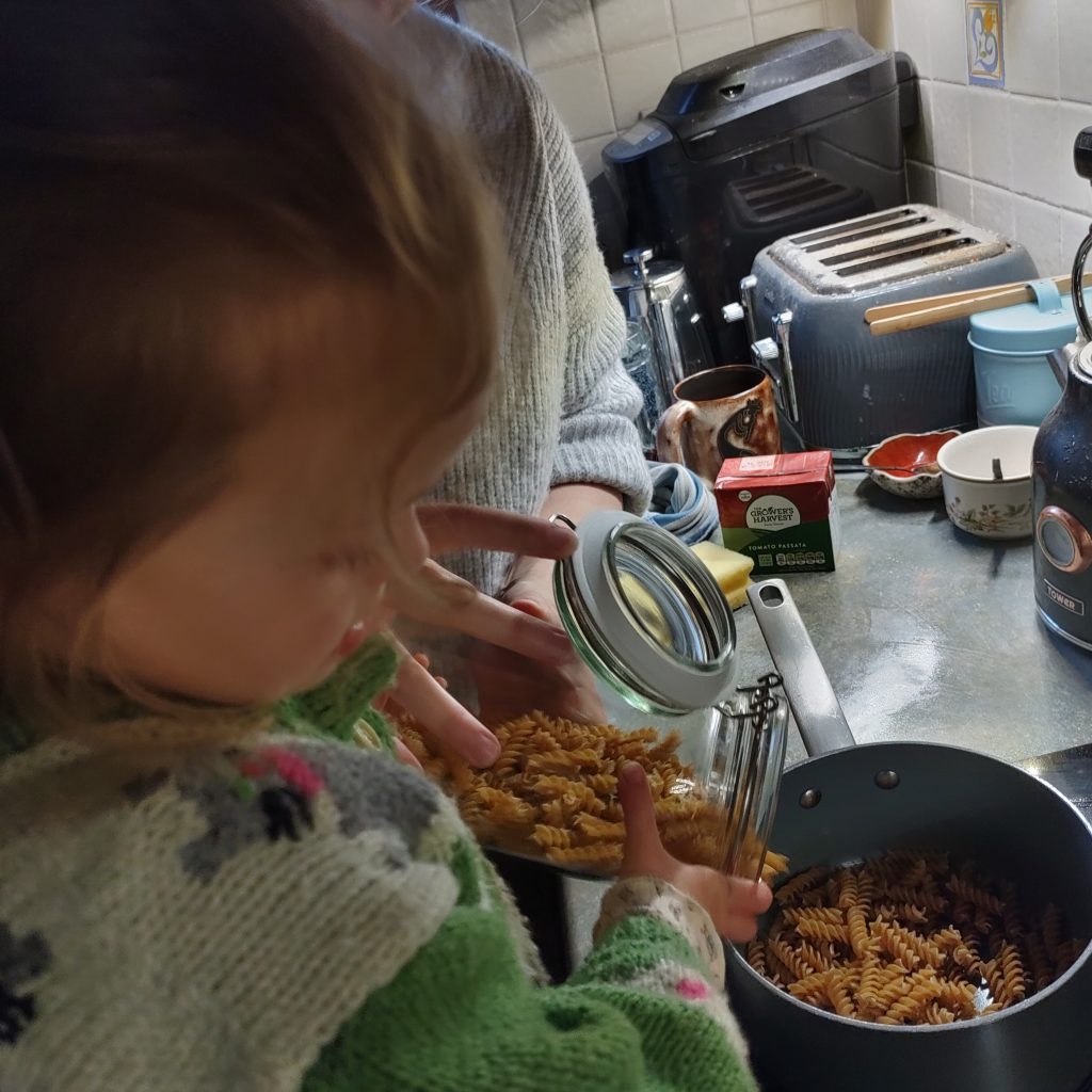 A little girl, with some help from her mother, pours some dried pasta into a pan.