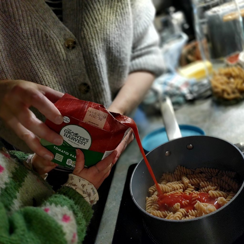 A little girl, with some help from her mum, adds some passata to the cooked pasta.