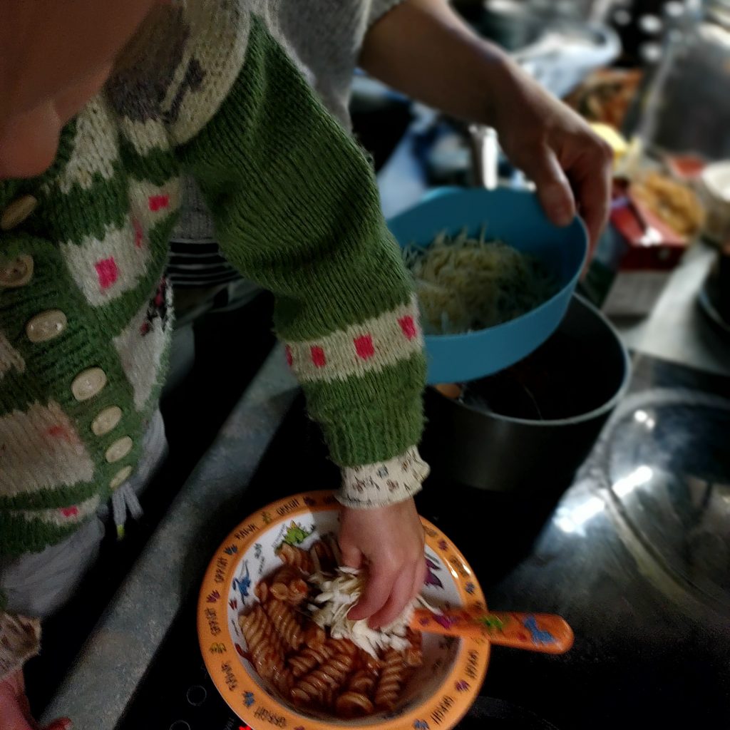 A little girl adds some cheddar cheese to a bowl of pasta and passata.