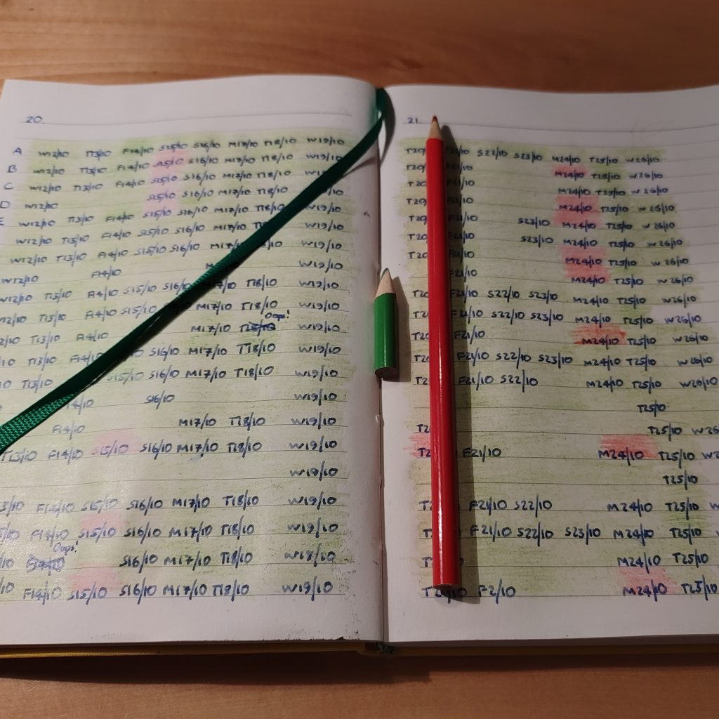 An open notebook, in which is written a series of dates, most shaded green but some shaded red. In the middle of the notebook lie two pencils: one very short green pencil, and one very long red pencil.