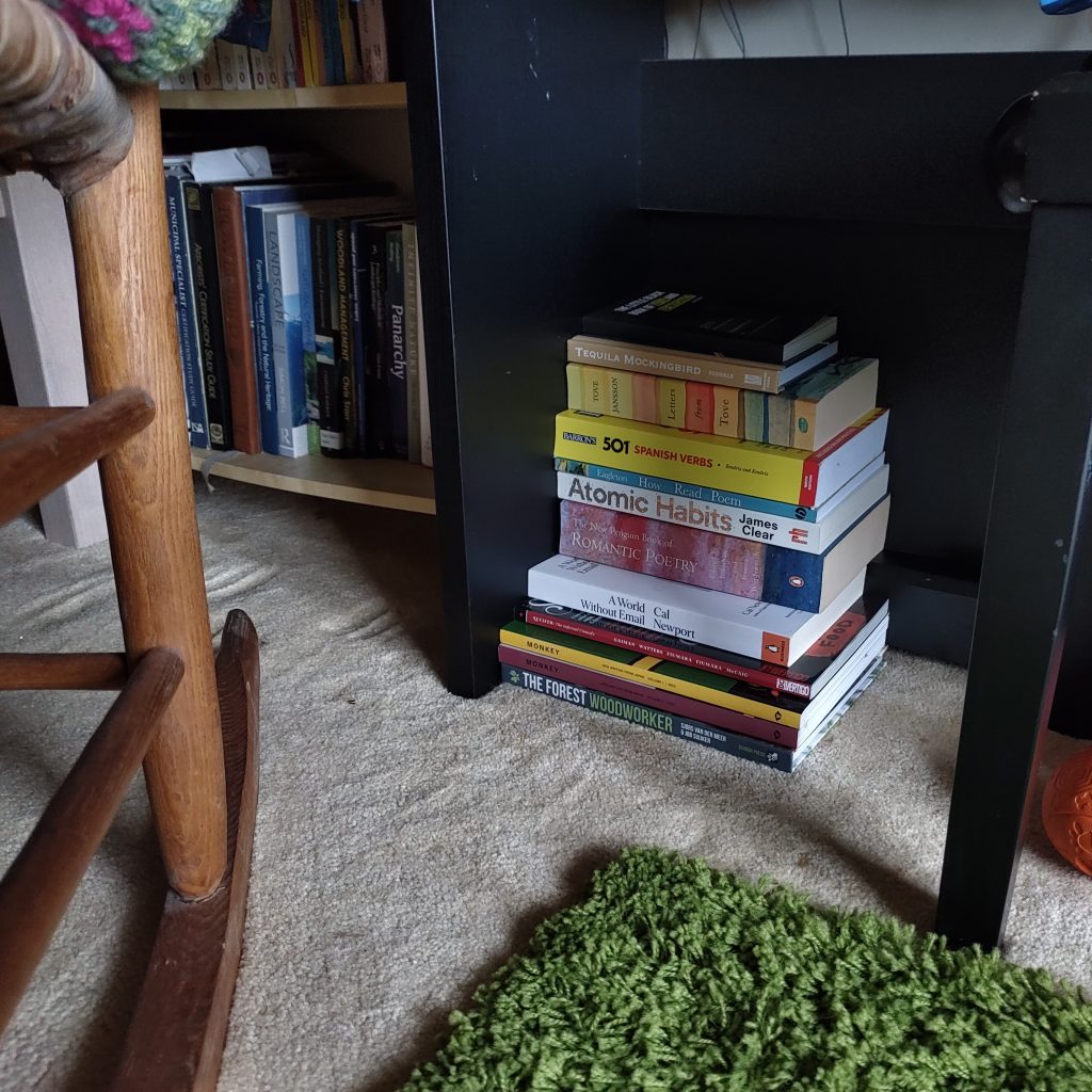 A pile of books is heaped underneath an upright electric piano. To the left, more bookcases, packed with books, can be seen stretching out of shot behind a rocking chair.